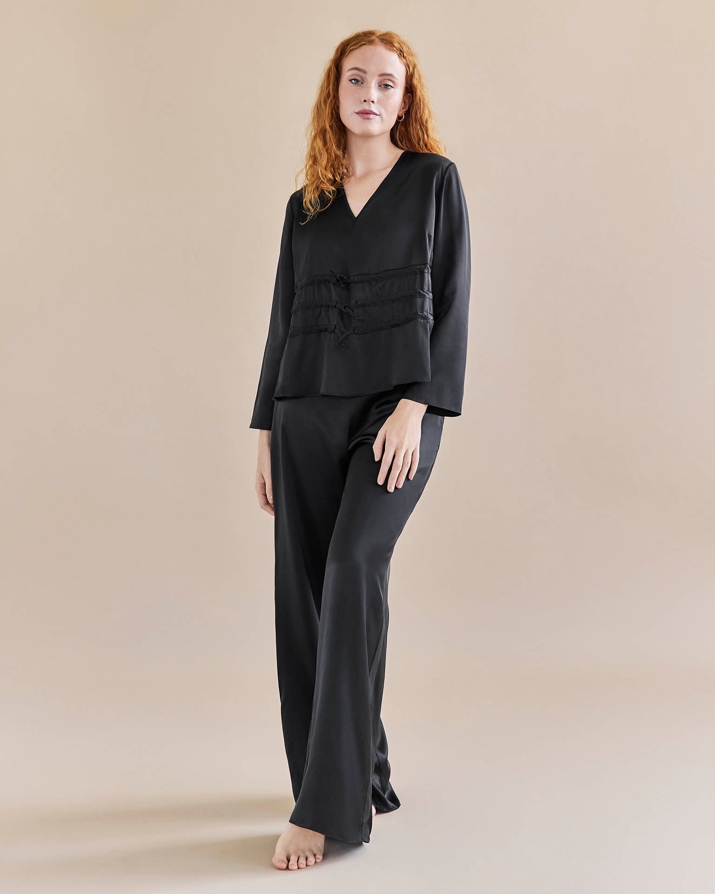 Model wears black silk tie waist top worn loose, and a pair of black, silk, wide legged trousers with a tuxedo stripe.