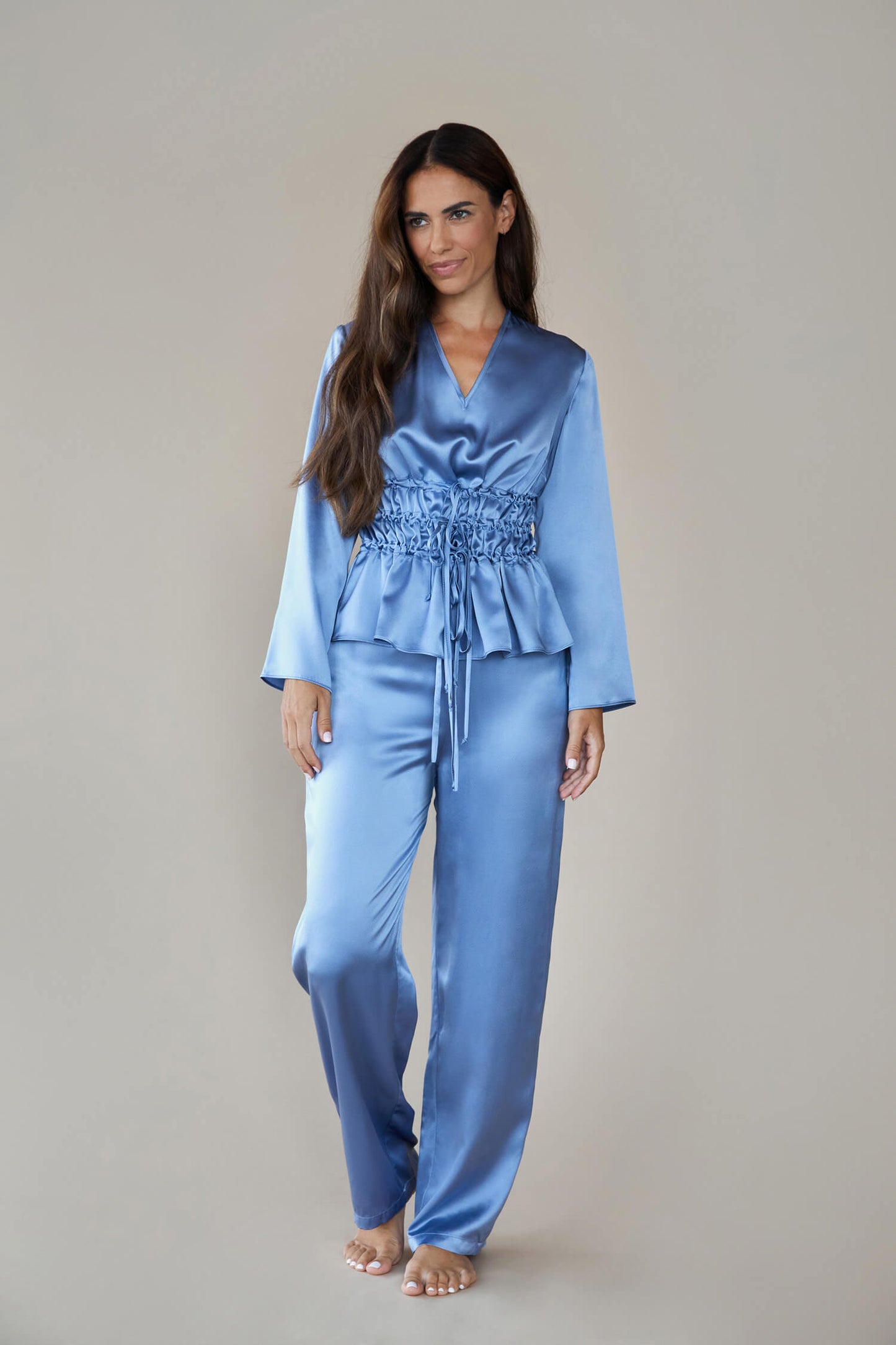 Model wears silk satin loungewear set in colour pewter sky (blue). Her top is a long sleeved, V neck top. It is shown with the waist ties untied to give a loose, easy fit rather than the corseted style waist. She wears straight legged pyjama style silk trousers with a drawstring, elasticated waist.