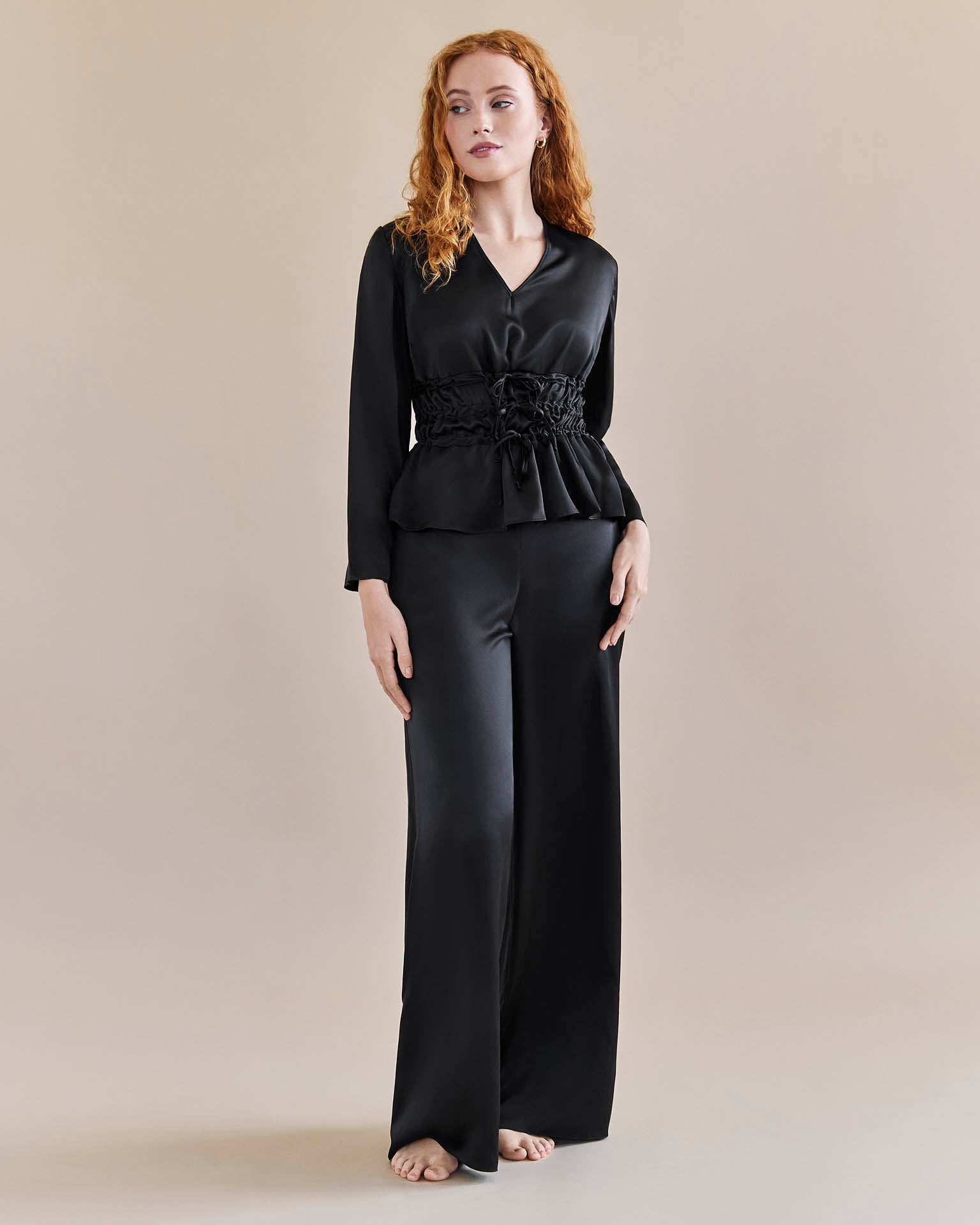 Model wears black silk tie waist top cinched in at the waist for an hourglass look and a pair of black, silk, wide legged trousers with a tuxedo stripe.