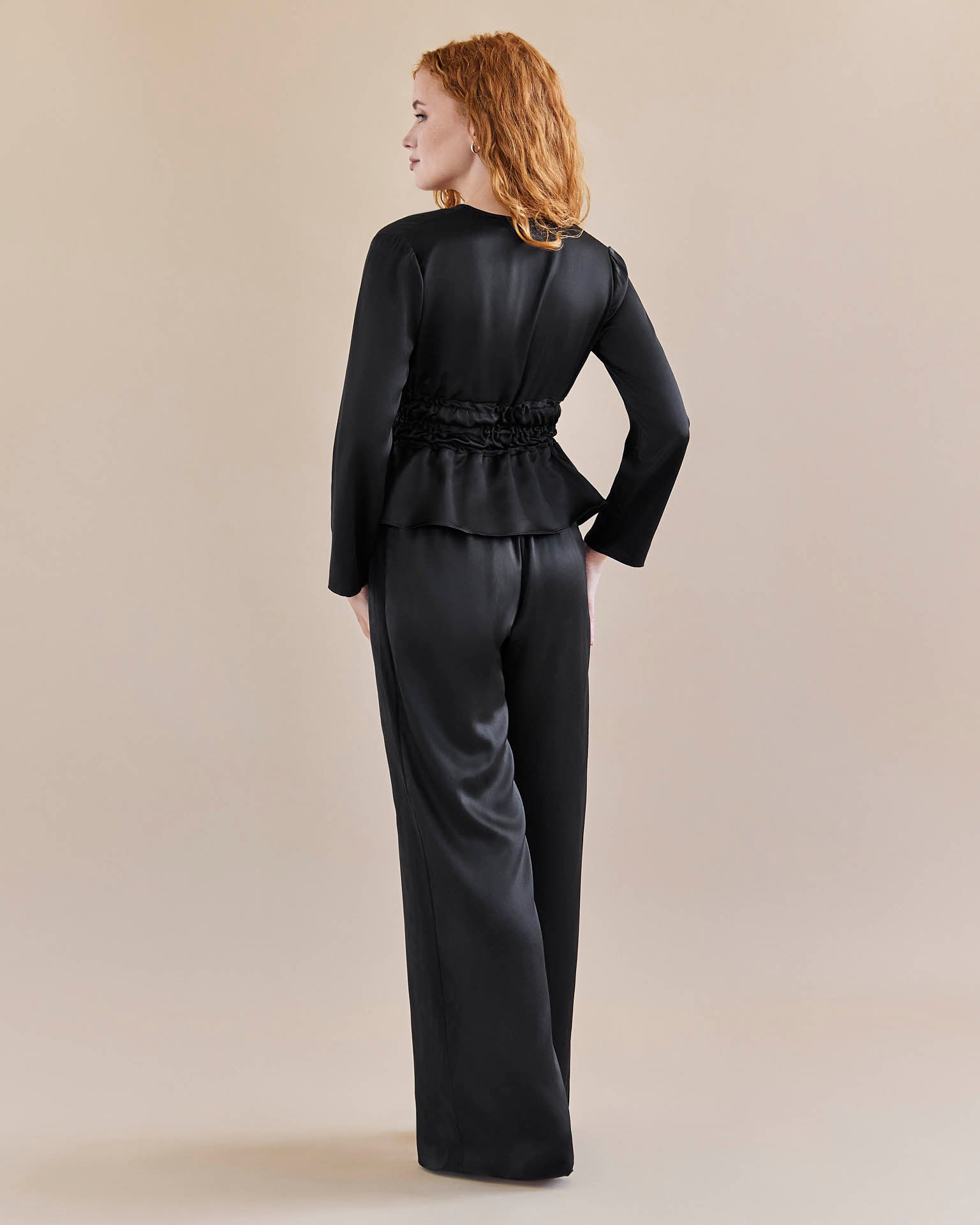 Model wears black silk tie waist top cinched in at the waist for an hourglass look and a pair of black, silk, wide legged trousers with a tuxedo stripe. Pictured from the back
