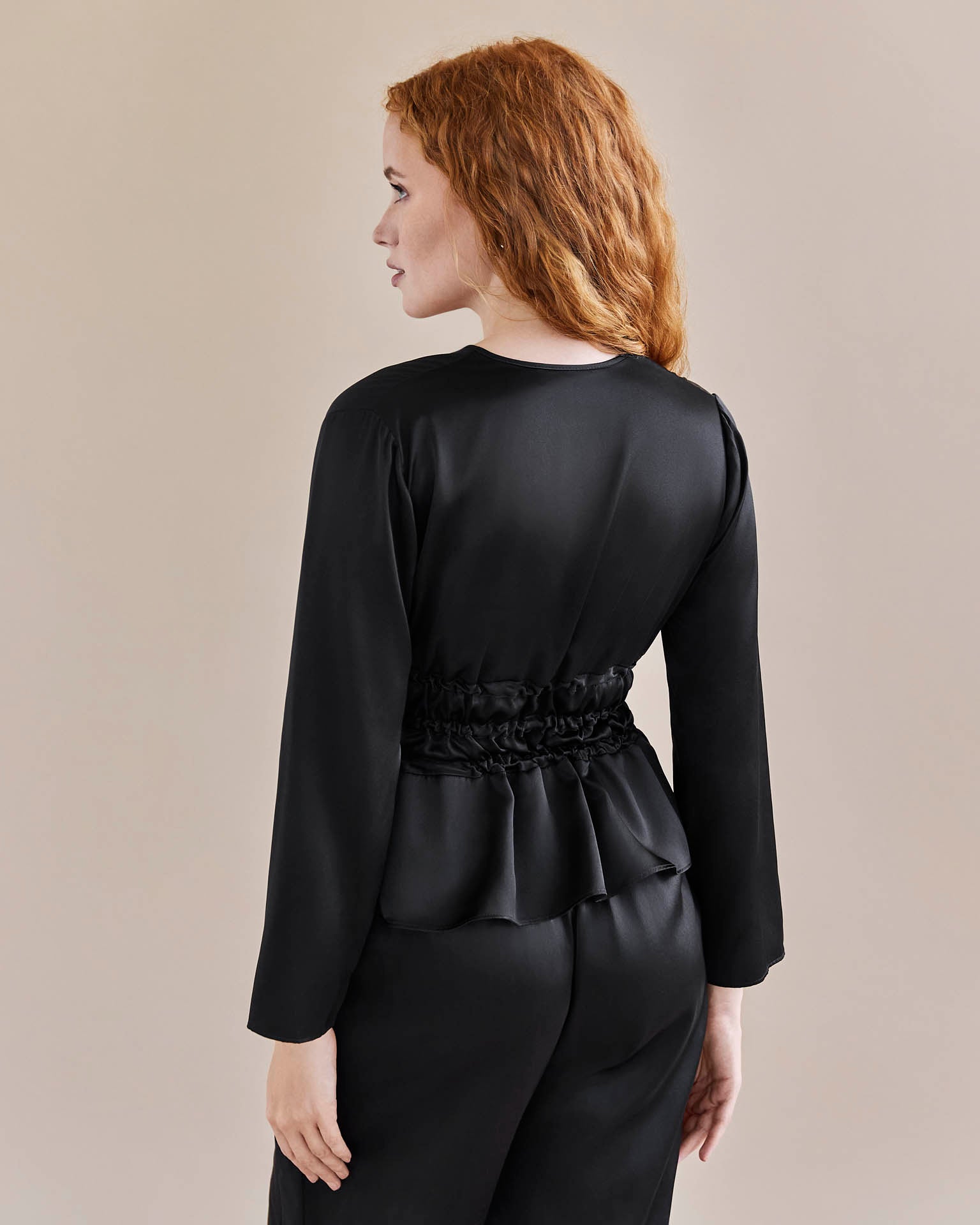 Detail shot Model wears black silk tie waist top cinched in at the waist for an hourglass look and a pair of black, silk, wide legged trousers with a tuxedo stripe. Pictured from the back