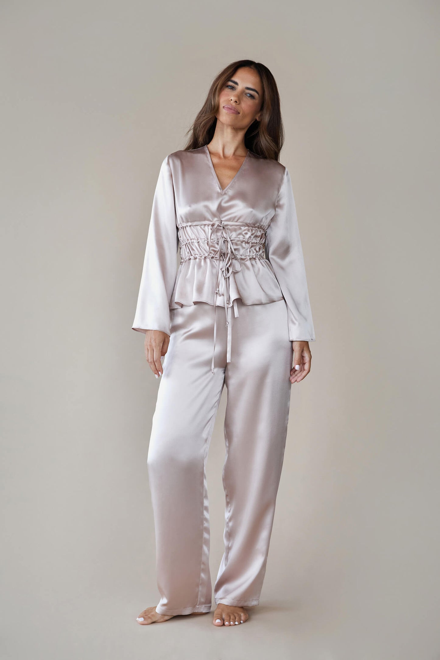 Model wears silk satin loungewear set in colour rose fawn. Her top is a long sleeved, V neck top with a corset style waist and she wears straight legged pyjama style silk trousers with a drawstring, elasticated waist. She looks towards the camera