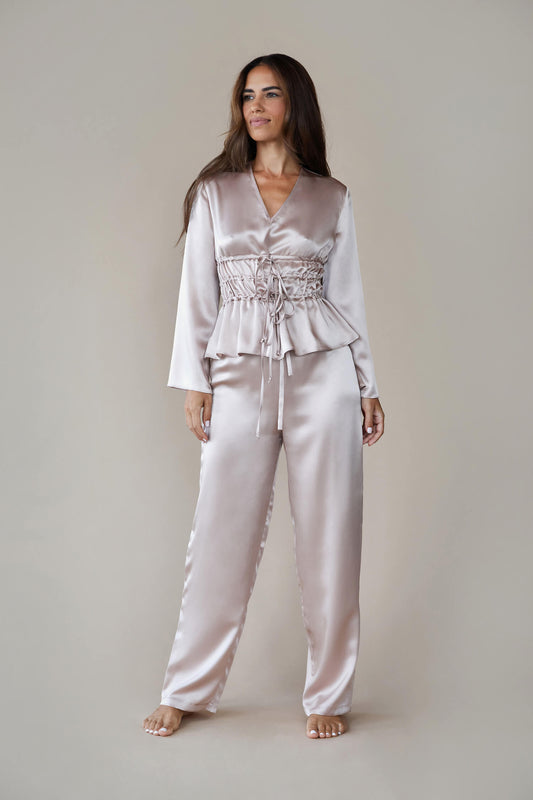 Model wears silk satin loungewear set in colour rose fawn. Her top is a long sleeved, V neck top with a corset style waist and she wears straight legged pyjama style silk trousers with a drawstring, elasticated waist. 