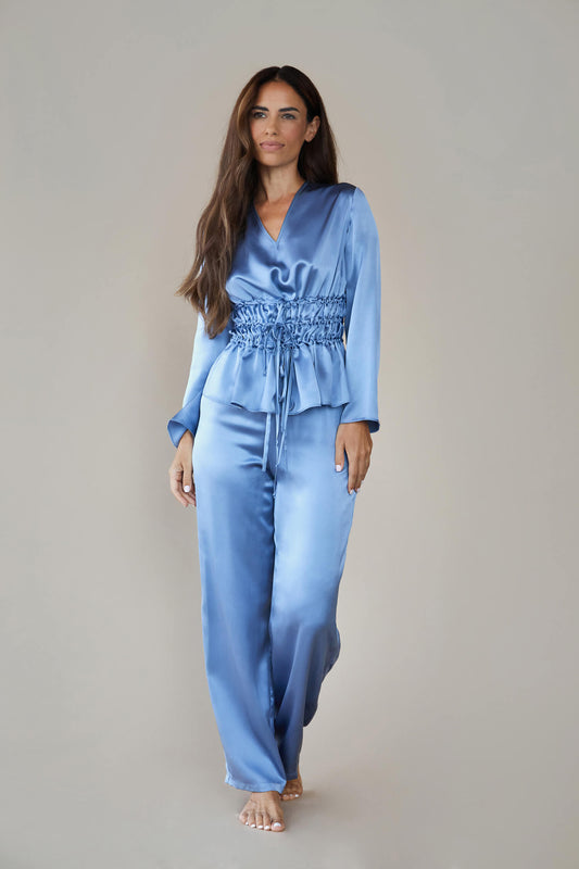 Model wears silk satin loungewear set in colour pewter sky (blue). Her top is a long sleeved, V neck top with a corset style waist and she wears straight legged pyjama style silk trousers with a drawstring, elasticated waist. 