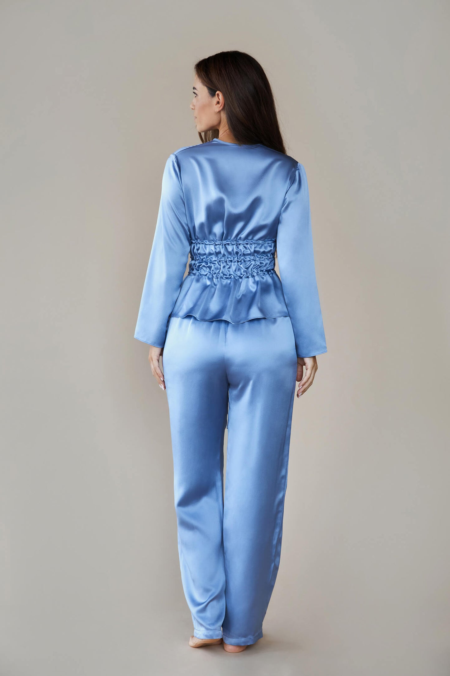 Shot from back to show the back of the set, the model wears silk satin loungewear set in colour pewter sky (blue). Her top is long sleeved with a corset style waist and she wears straight legged pyjama style silk trousers with a drawstring, elasticated waist.