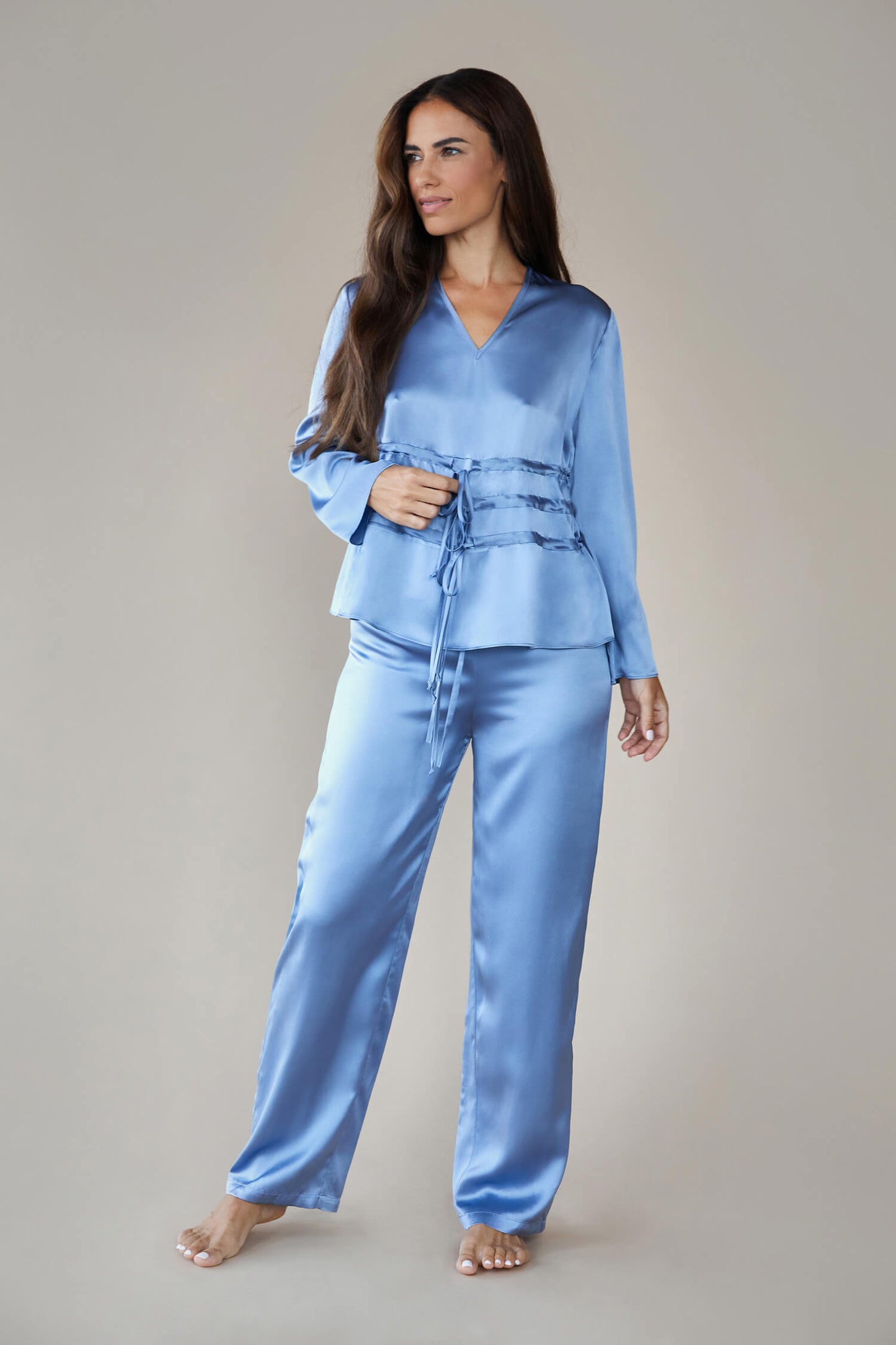 Model wears silk satin loungewear set in colour pewter sky (blue). Her top is a long sleeved, V neck top. It is shown with the waist ties untied to give a loose, easy fit rather than the corseted style waist. She wears straight legged pyjama style silk trousers with a drawstring, elasticated waist. 