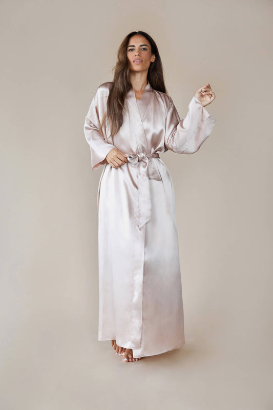 Model wears kimono style luxury silk robe in colour rose fawn tied at the waist. She looks at the camera confidently and raises her hand, showing the wider sleeve
