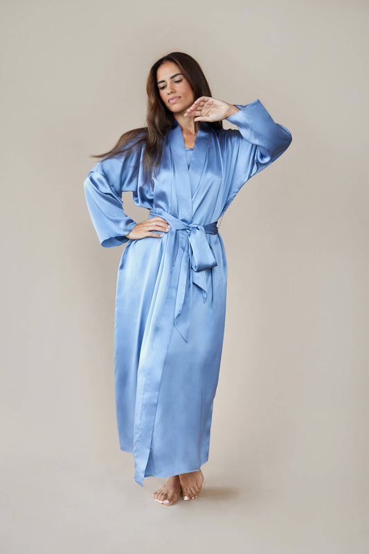 Model wears kimono style luxury silk robe in blue tied at the waist. She looks down wistfully and raises her hand to her face