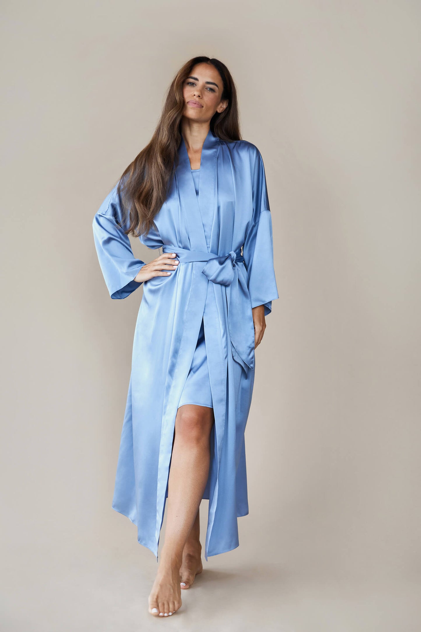 Model wears a  kimono style luxury silk robe In blue tied at the waist using a bow. She looks to the camera confidently with one hand on her hip, revealing a hint of a silk dress underneath, with one leg rested in front of the other