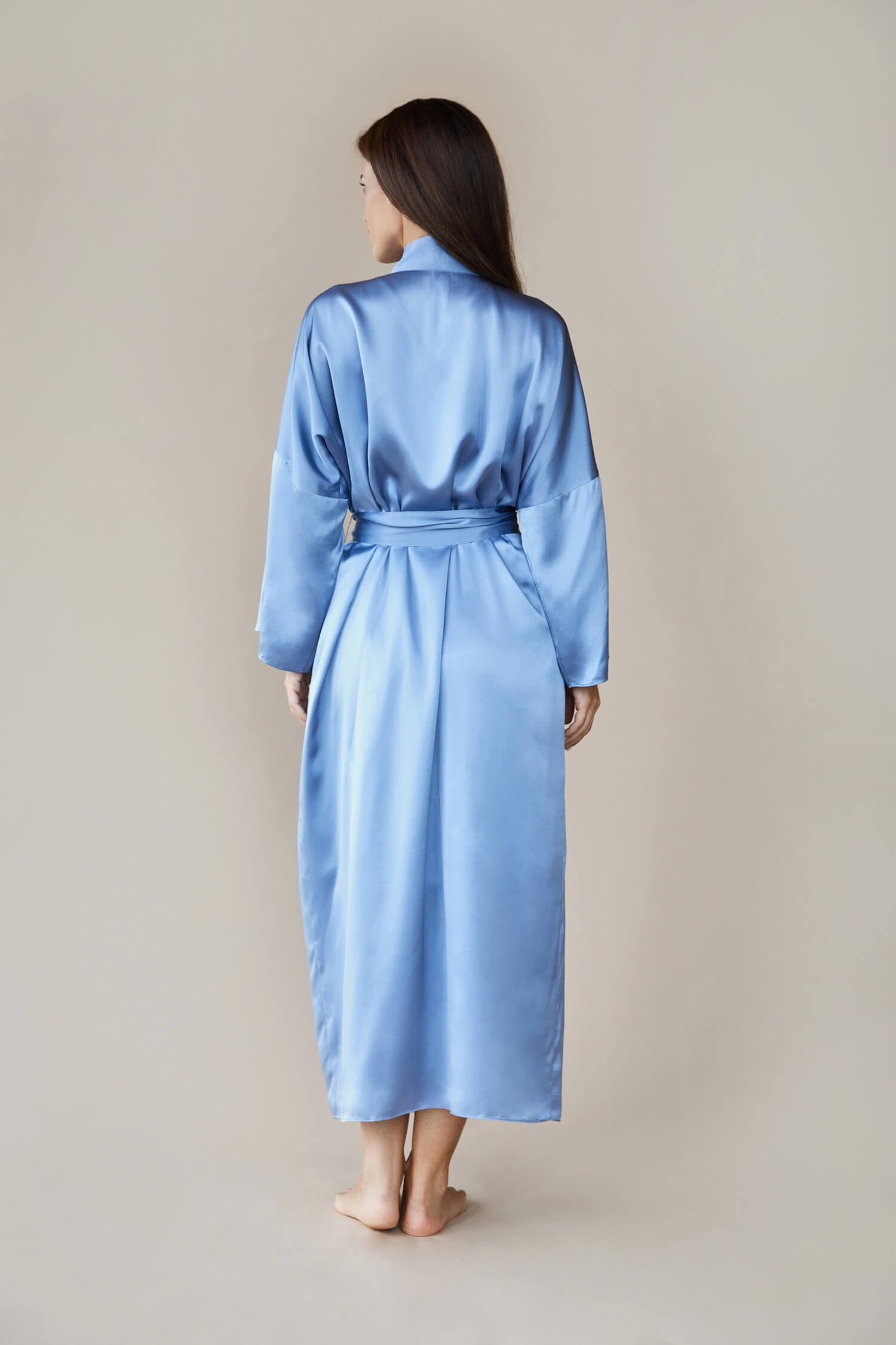 Model shot from the back wearing a kimono style luxury silk robe In blue tied at the waist. She looks to the side with her hands by her side.