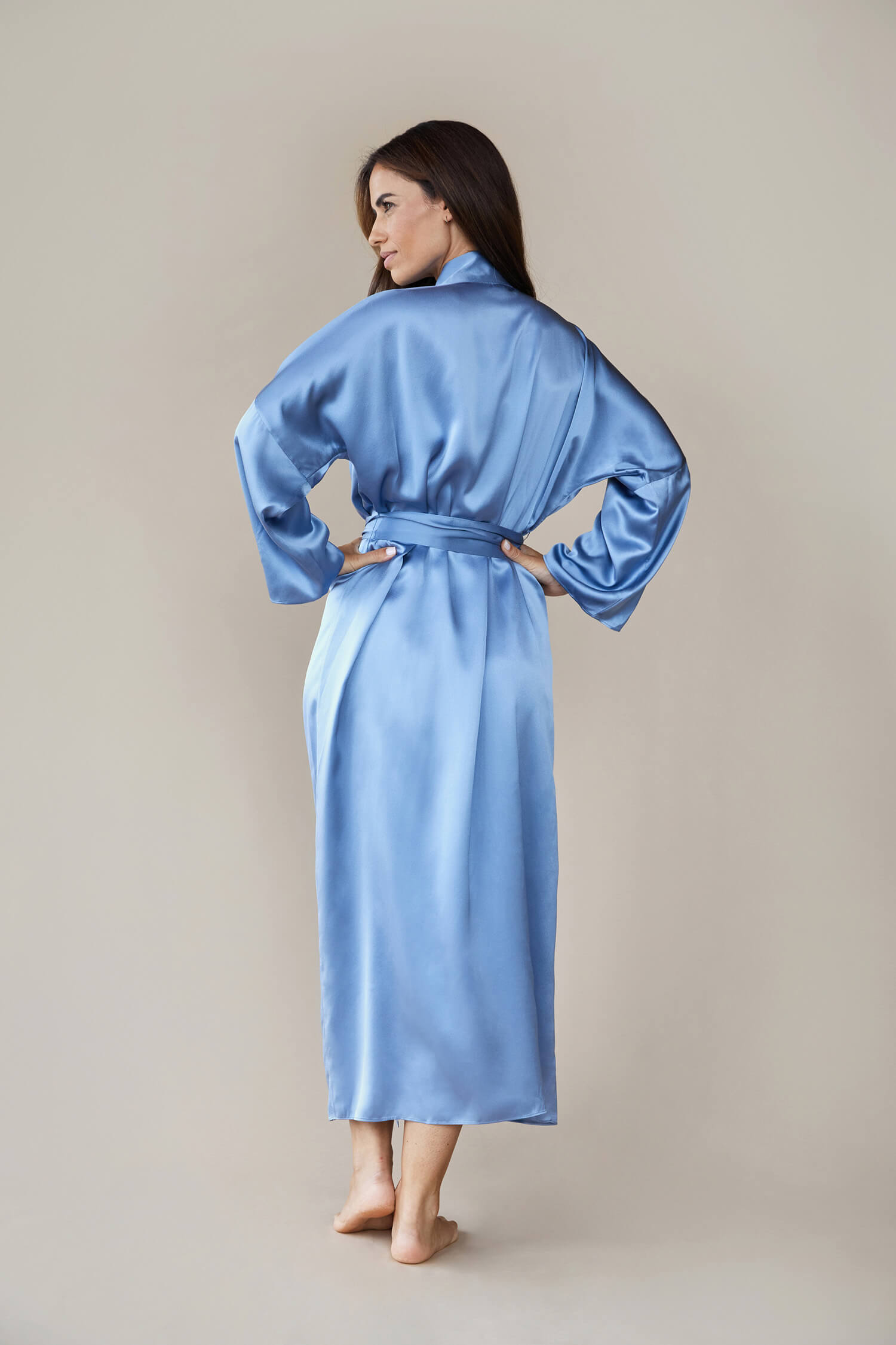 Model shot from the back wearing a  kimono style luxury silk robe In blue tied at the waist. She looks to the side with her hands on her waist elegantly