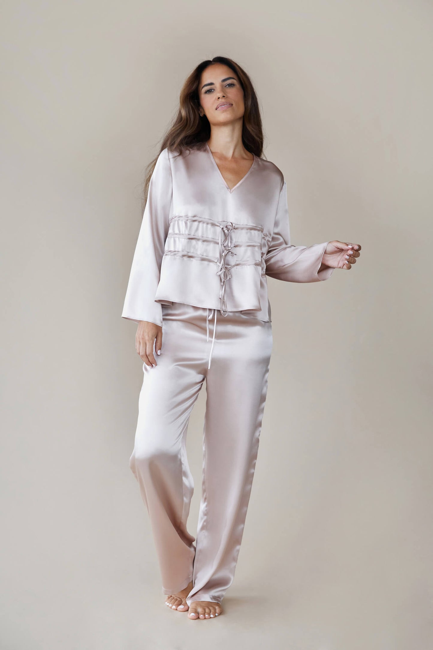  Model wears silk satin loungewear set in colour rose fawn. Her top is a long sleeved, V neck top. It is shown with the waist ties untied to give a loose, easy fit rather than the corseted style waist. She wears straight legged pyjama style silk trousers with a drawstring, elasticated waist. 